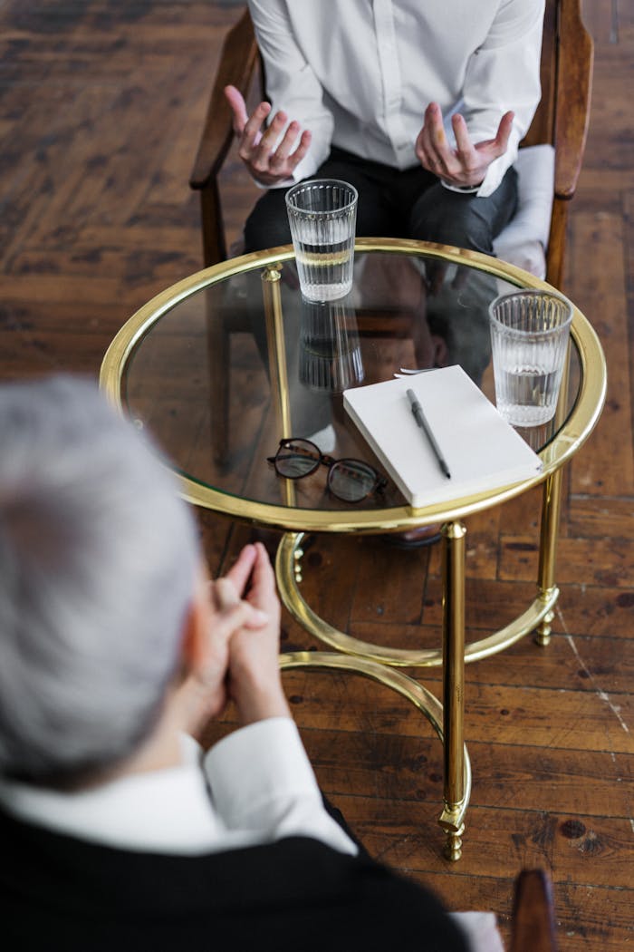 Servizi Two adults discussing mental health in a counseling session across a glass table indoors.